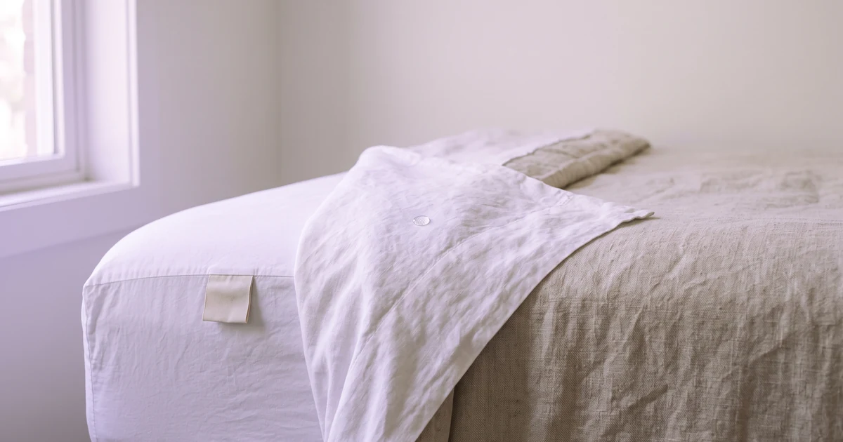 Corner of a made bed with white sheets folded back and a blank fabric care tag visible, illustrating that bedding labels do not disclose PFAS finishes